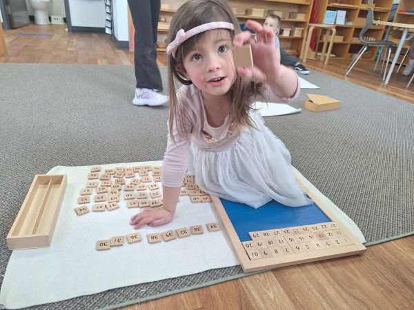 Casa student kneeling on a mat on the floor holding a small wooden tile from Montessori material. 