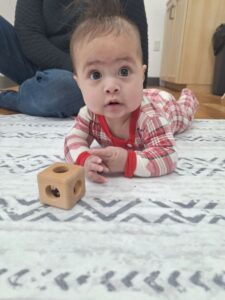 Infant lying on a grey and white rug playing with wooden material.