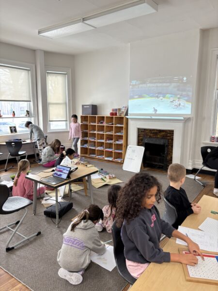 Children working in their classroom with the Olympics projected on the wall over a fireplace.