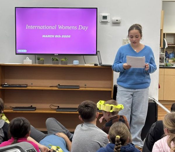 A Upper El student presents to her classmates in front of a TV screen that reads International Women's Day.