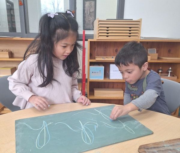 Two students writing cursive letters on a green chalk board on a table. 