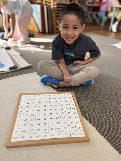 A Casa child sits on the floor in front of a completed Montessori 100 Board.
