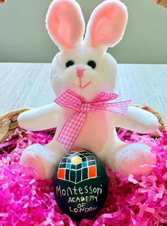 Photo of a stuffed easter bunny in a basket with an egg decorated with the school's trinomial cube logo and the words Montessori Academy of London. 