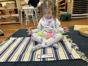 A girl sits on a striped mat with crossed legs, meditating during the Montessori Silence Game.